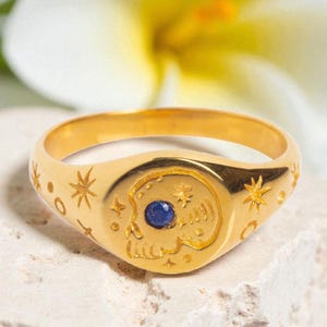 May include: A gold-toned signet ring featuring an engraved skull motif on the face, accented with a small, round, deep blue gemstone set within the skull. The band is decorated with engraved star and circular patterns surrounding the central design.
