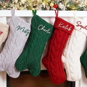 Puede incluir: Una fila de seis calcetines navideños de punto trenzado colgados de una repisa blanca. Los calcetines son beige, gris claro, verde oscuro, rojo y blanco, cada uno con un nombre personalizado bordado en letra cursiva: Roxy, Misty, Charlie, Kaleb y Sophie.