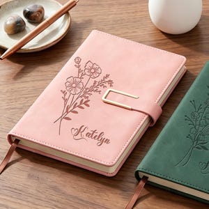 May include: Two personalized journals, one pink and one green, with floral designs and gold-colored clasps. The pink journal has the name "Katelyn" and the green journal has the name "Mckayla" engraved on the cover. Both have a ribbon bookmark.