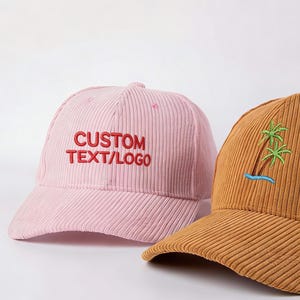Peut inclure: Deux casquettes de baseball en velours côtelé. L'une est rose clair avec "CUSTOM TEXT/LOGO" brodé en rouge. L'autre est marron avec une broderie de palmier et d'océan en vert et bleu.