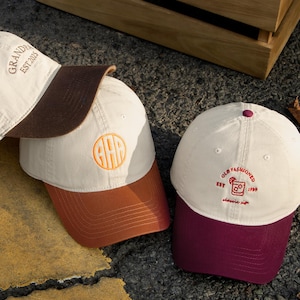 May include: Three baseball caps with different designs. One is tan with a brown brim and the text "GRANDPA EST 2015". Another is tan with an orange brim and an embroidered monogram. The third is white with a burgundy brim and "OLD FASHIONED" text.