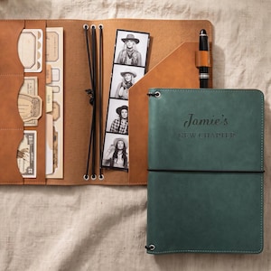 May include: A tan leather-like journal cover with interior pockets containing paper ephemera and a photo strip, paired with a dark green journal insert featuring the text Jamie's NEW CHAPTER and a black elastic band closure with a black pen.