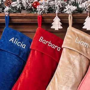 Op de afbeelding: Drie kerstsokken van fluweel in blauw, rood en champagne goud, elk met een omgeslagen boord en een naam geborduurd in wit garen. De sokken zijn gelabeld met Alicia, Barbara en Liam en hangen aan een houten schoorsteenmantel.