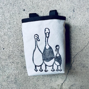 May include: A white canvas bag with a black top and handles. The bag features a black and white illustration of three geese. The geese are standing in a row, with the middle goose being the largest.