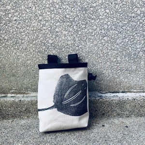 May include: A white canvas chalk bag with a black silhouette of a stingray printed on the front. The bag has black straps and a drawstring closure.