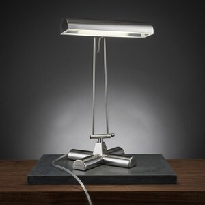 May include: A silver-toned metal desk lamp featuring a rectangular horizontal shade, two thin vertical support rods, and a cross-shaped base. The lamp emits a bright light from beneath the shade and includes a clear power cord.