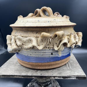 May include: A round ceramic lidded casserole dish with a speckled tan glaze, featuring a ruffled clay trim around the center and a blue band near the base. The lid has a looped handle, and the word REECE is stamped on the lower side.