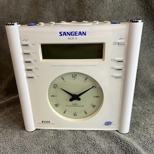 May include: A white Sangean RCR-3 FM/AM clock radio with a digital display and an analog clock face. The device features multiple control buttons labeled Time, Date, Nap, and alarm settings, with the text HWS Humane Waking System printed below the clock hands.