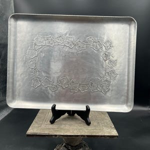 May include: A rectangular silver-toned metal serving tray featuring a hammered finish and a central embossed border design of ivy leaves and vines. The tray has rounded corners and a slightly raised edge, showcasing a metallic sheen.