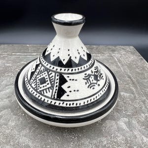 May include: A ceramic tagine with a conical lid and a wide base. The piece features a white glazed finish with a black hand-painted geometric pattern and a subtle crackle texture. The rim of the lid and the base are accented with a solid black band.