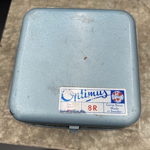 Könnte beinhalten: Ein hellblauer Metallkoffer für einen Optimus 8R Campingkocher. Auf der Oberseite befindet sich ein rechteckiger weißer Aufkleber mit blauem und rotem Text, der Optimus, 8 R, For gasoline und Camp Stove Made in Sweden sowie ein Globus-Logo zeigt.