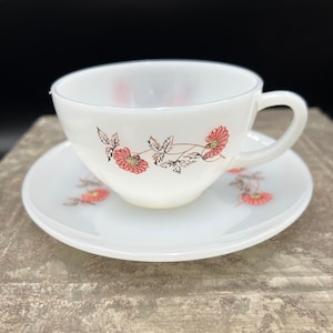 May include: White teacup and saucer set with a floral design. The cup and saucer are decorated with pink flowers and brown stems. The cup has a handle and sits on a matching saucer.