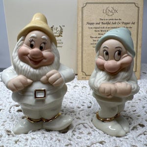 May include: A Lenox Happy and Bashful salt and pepper set featuring two porcelain dwarf figurines with white clothing, gold accents, and painted facial features. The set includes a certificate of authenticity with the text Happy and Bashful Salt & Pepper Set.