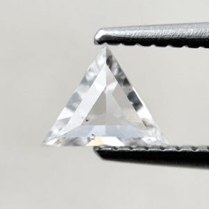 Op de afbeelding: Een heldere, driehoekig geslepen edelsteen, vastgehouden door een pincet. De edelsteen heeft een gefacetteerd oppervlak dat licht reflecteert. De vorm is een perfecte gelijkzijdige driehoek, die de helderheid en precisie van de slijping laat zien.