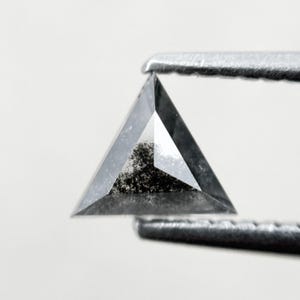 May include: A triangular-shaped, gray diamond held by metal tweezers. The diamond has a unique, speckled interior and a polished, faceted surface. The gem is set against a plain white background, highlighting its shape and clarity.