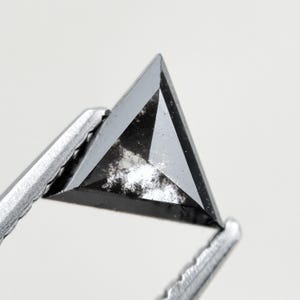 Puede incluir: Primer plano de un diamante negro triangular, sujeto con pinzas plateadas. El diamante presenta un patrón moteado con áreas claras y oscuras. Los bordes son afilados y bien definidos. Una gema única.