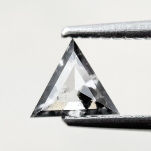 Könnte beinhalten: Ein dreieckig geformter, facettierter Diamant, der von einer Pinzette gehalten wird. Der Diamant ist überwiegend grau mit schwarzen Einschlüssen und einer reflektierenden Oberfläche. Der Edelstein hat eine einzigartige Form, ideal für Schmuck.