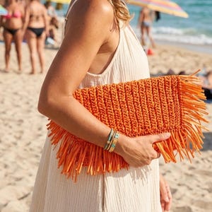 May include: An orange clutch with fringe detail. The rectangular bag is made of a woven material and is held by a person on a beach. The clutch has a textured appearance and a bright, solid color.