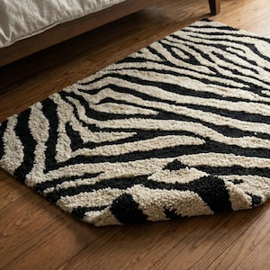 以下が含まれることがあります： 黒と白のゼブラ柄のラグで、丸みを帯びたエッジがあります。 ラグは柔らかく、質感のある素材でできており、木製の床に置かれています。 ラグは木製のベッドフレームの下に部分的にあります。