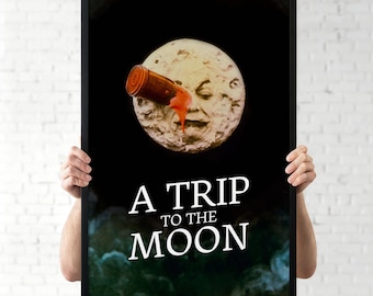 Puede incluir: Póster enmarcado en negro con el texto "A Trip to the Moon". El póster muestra una ilustración vintage de la luna con una cara, un cañón y líquido rojo saliendo del cañón.