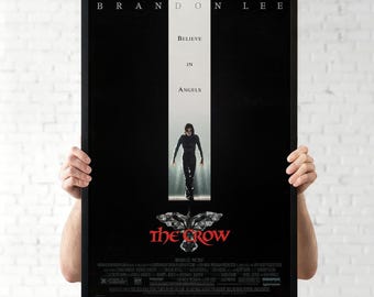 May include: A framed movie poster for "The Crow" featuring Brandon Lee. The poster has a black background with the text "Believe in Angels" and the movie title in red. A figure in black stands in a white vertical stripe.