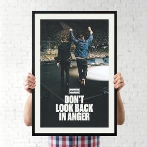 May include: A framed poster featuring a photograph of two individuals standing on a concert stage facing a large crowd. The text "oasis" appears in a small box above the bold, white, capitalized text "DON'T LOOK BACK IN ANGER" at the bottom.