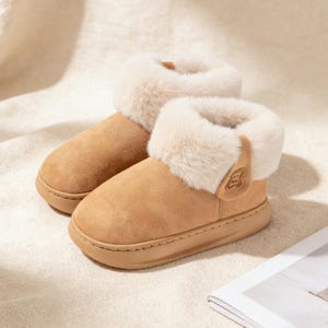May include: A pair of tan-colored, ankle-high slippers with a plush, cream-colored faux fur trim. The slippers have a rounded toe and a small, decorative emblem on the side. The soles are a matching tan color.