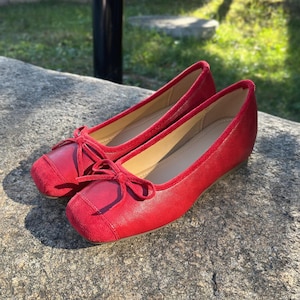 May include: A pair of red ballet flats with a square toe and a small bow detail. The shoes are made of a smooth material with a suede toe cap. The shoes are sitting on a stone surface.
