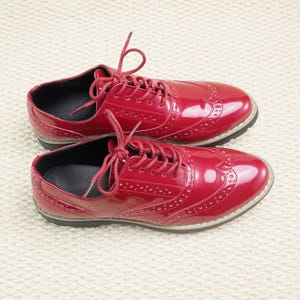 Peut inclure: Une paire de chaussures Oxford rouges brillantes avec des perforations d&eacute;coratives et des lacets rouges assortis. Les chaussures ont un design classique &agrave; bout d'aile et une semelle noire. Elles sont pos&eacute;es sur une surface blanche textur&eacute;e.
