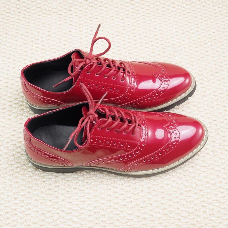 Peut inclure: Une paire de chaussures Oxford rouges brillantes avec des perforations d&eacute;coratives et des lacets rouges assortis. Les chaussures ont un design classique &agrave; bout d'aile et une semelle noire. Elles sont pos&eacute;es sur une surface blanche textur&eacute;e.