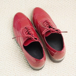 Peut inclure: Une paire de chaussures Oxford rouges brillantes avec des perforations d&eacute;coratives et des lacets rouges assortis. Les chaussures sont pr&eacute;sent&eacute;es sur une surface textur&eacute;e blanc cass&eacute;. Design classique.