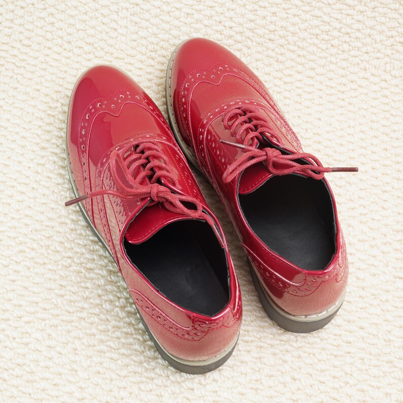 Peut inclure: Une paire de chaussures Oxford rouges brillantes avec des perforations d&eacute;coratives et des lacets rouges assortis. Les chaussures sont pr&eacute;sent&eacute;es sur une surface textur&eacute;e blanc cass&eacute;. Design classique.