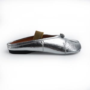 May include: A silver metallic slip-on shoe with a small bow detail and a tan elastic strap. The shoe has a black sole and a brown interior. The shoe is designed for comfort and style.