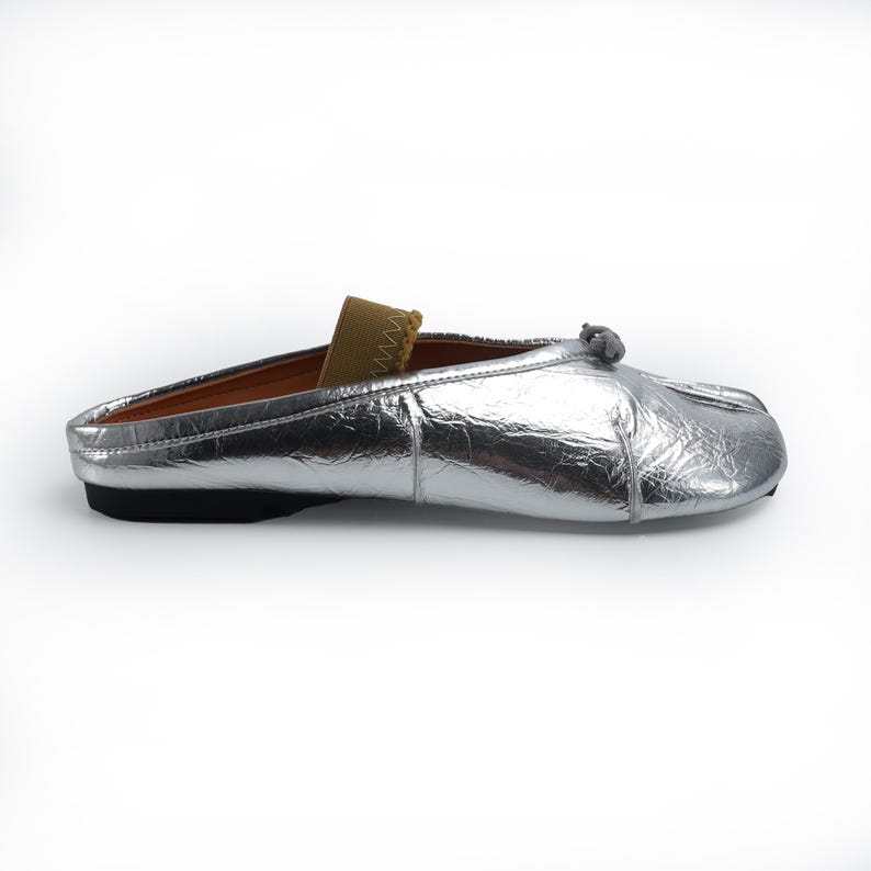 May include: A silver metallic slip-on shoe with a small bow detail and a tan elastic strap. The shoe has a black sole and a brown interior. The shoe is designed for comfort and style.