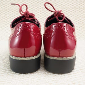 Peut inclure: Paire de chaussures &agrave; plateforme rouges brillantes avec des perforations d&eacute;coratives. Les chaussures ont un style Oxford classique avec des lacets rouges et une semelle noire &eacute;paisse. Les chaussures sont pr&eacute;sent&eacute;es sur une surface textur&eacute;e blanche.