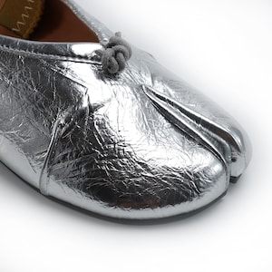 May include: Silver metallic split-toe ballet flat. The shoe features a textured, crinkled appearance and a small knot detail on the top. The sole is a contrasting tan color. The shoe is designed with a split toe, separating the big toe from the other toes.