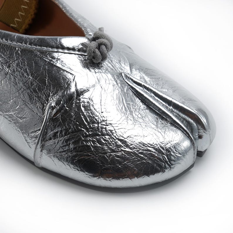 May include: Silver metallic split-toe ballet flat. The shoe features a textured, crinkled appearance and a small knot detail on the top. The sole is a contrasting tan color. The shoe is designed with a split toe, separating the big toe from the other toes.