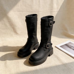 May include: A pair of black knee-high boots with a chunky sole. Each boot has a buckle and strap detail around the ankle and calf. The boots are made of a smooth, matte material.