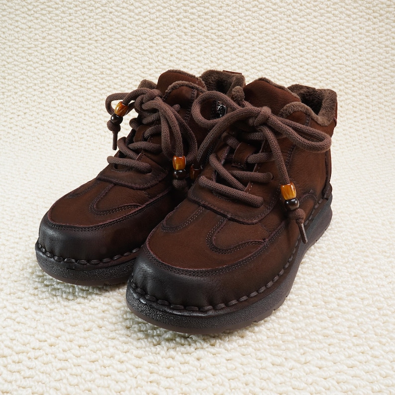 May include: A pair of brown leather ankle boots with dark brown soles and laces. The boots have a rounded toe and are lined with a soft, light brown material. The laces are secured with small wooden beads.
