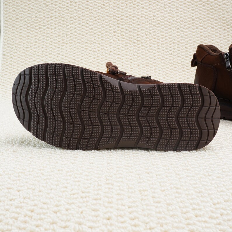 May include: Close-up of the sole of a brown leather boot. The sole has a textured, wavy pattern for grip. The boot is on a white, textured surface.
