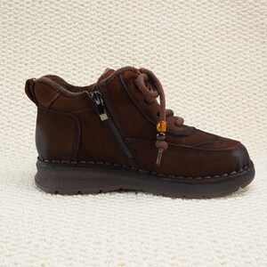 May include: Brown leather ankle boot with a side zipper and lace-up closure. The boot features a rounded toe, a low heel, and decorative stitching. A small beaded accent hangs from the laces. The boot is designed for comfort and style.