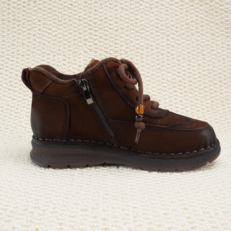 May include: Brown leather ankle boot with a side zipper and lace-up closure. The boot features a rounded toe, a low heel, and decorative stitching. A small beaded accent hangs from the laces. The boot is designed for comfort and style.