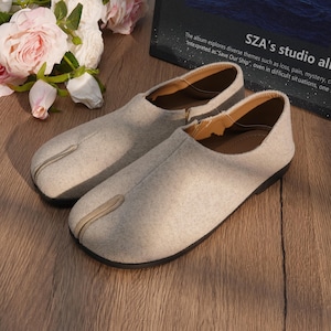 Puede incluir: Un par de zapatos grises claros con puntera dividida y suela negra. Los zapatos tienen una parte superior de tela texturizada y un ribete en contraste de color marrón claro. Zapatos cómodos e informales.