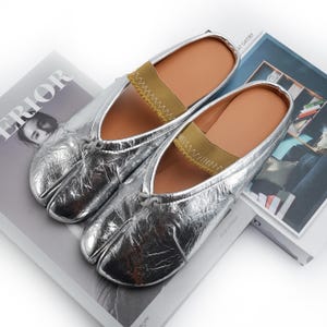 May include: A pair of silver split-toe slippers with a metallic finish. The slippers have a brown insole and a tan elastic strap across the top. The shoes are resting on top of a magazine.