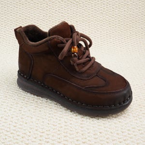 May include: Brown ankle boot with a lace-up design and a decorative bead on the laces. The boot features a padded collar and a stitched sole. The shoe is made of a suede-like material and has a dark brown sole.