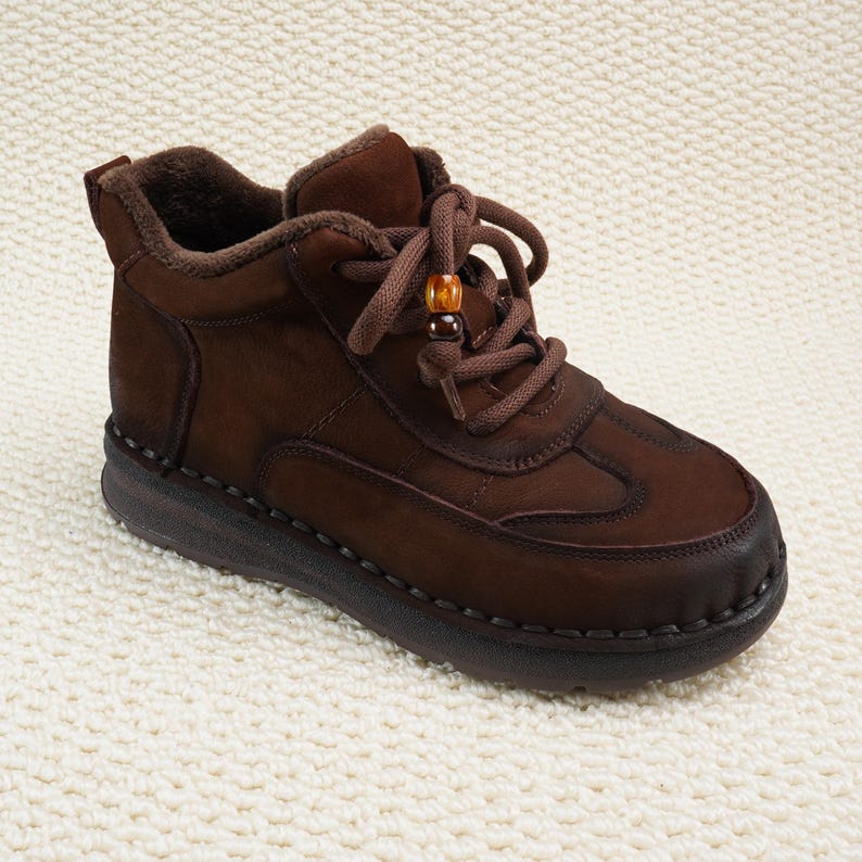May include: Brown ankle boot with a lace-up design and a decorative bead on the laces. The boot features a padded collar and a stitched sole. The shoe is made of a suede-like material and has a dark brown sole.