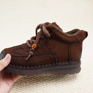 May include: Brown leather ankle boot with a rounded toe and lace-up closure. The boot features decorative stitching and a small wooden bead detail on the laces. The sole is a darker brown color.
