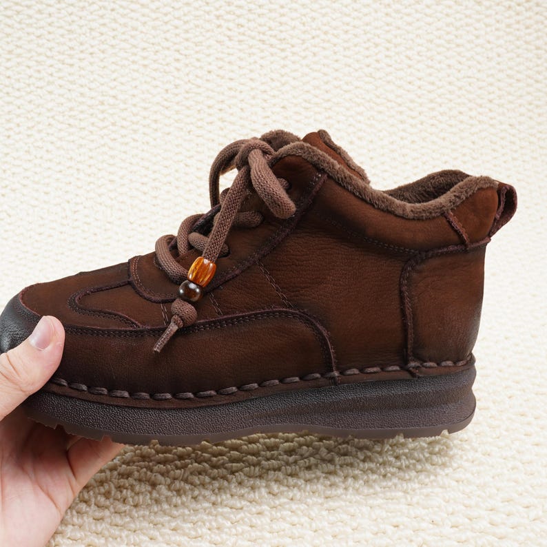 May include: Brown leather ankle boot with a rounded toe and lace-up closure. The boot features decorative stitching and a small wooden bead detail on the laces. The sole is a darker brown color.