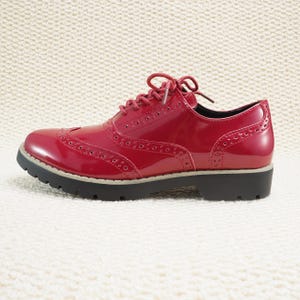 Peut inclure: Une paire de chaussures Oxford en cuir verni rouge avec un design brogue classique. Les chaussures ont une finition brillante, une semelle noire et des lacets rouges. Les chaussures mesurent environ 25 cm de long.