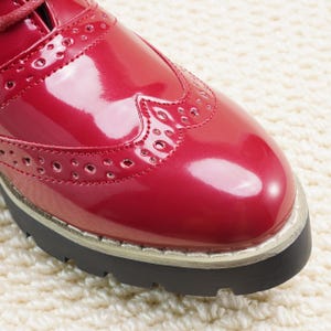 Peut inclure: Gros plan d'une chaussure oxford rouge brillant. La chaussure pr&eacute;sente des perforations d&eacute;coratives et une semelle &eacute;paisse noire. Le design de la chaussure comprend un style richelieu avec un bout rond et une fermeture &agrave; lacets.